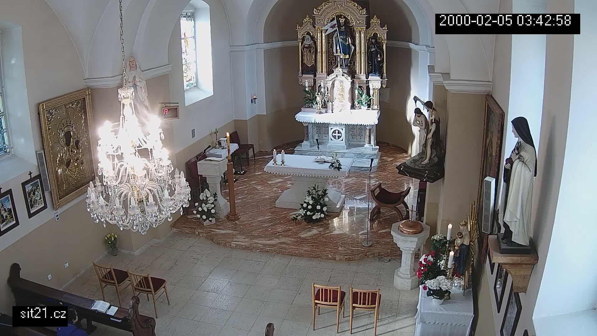 A church interior with a large chandelier, marble floors, an altar with floral arrangements, and several religious statues is brightly lit by sunlight streaming through a window.