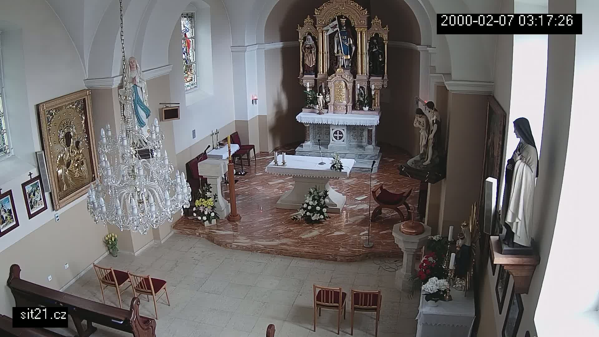 A church interior is shown, featuring an altar, several religious statues, a large crystal chandelier, and a few chairs arranged in the center.