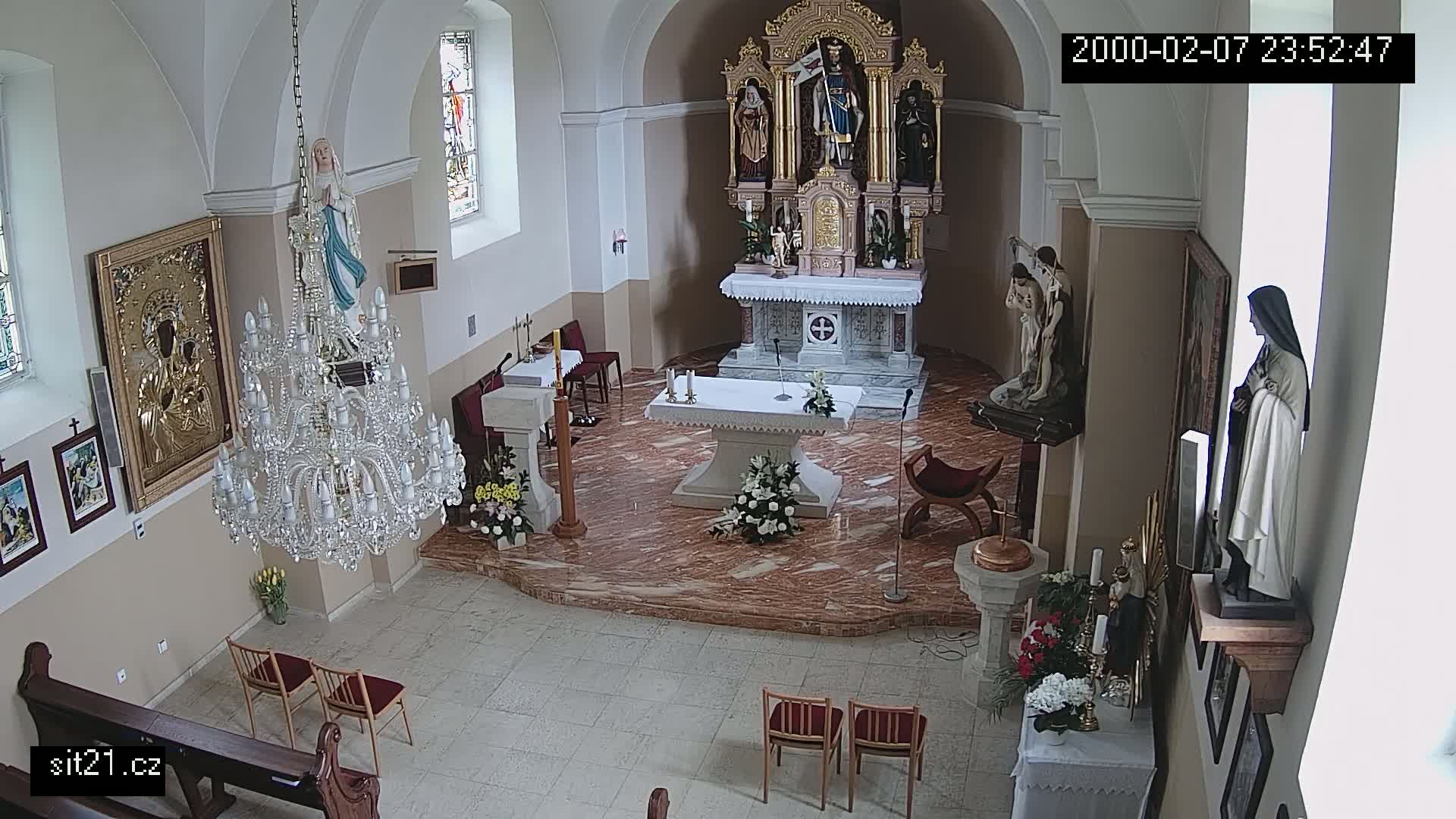 An indoor view of a church sanctuary shows an altar, statues, a large crystal chandelier, and several floral arrangements.