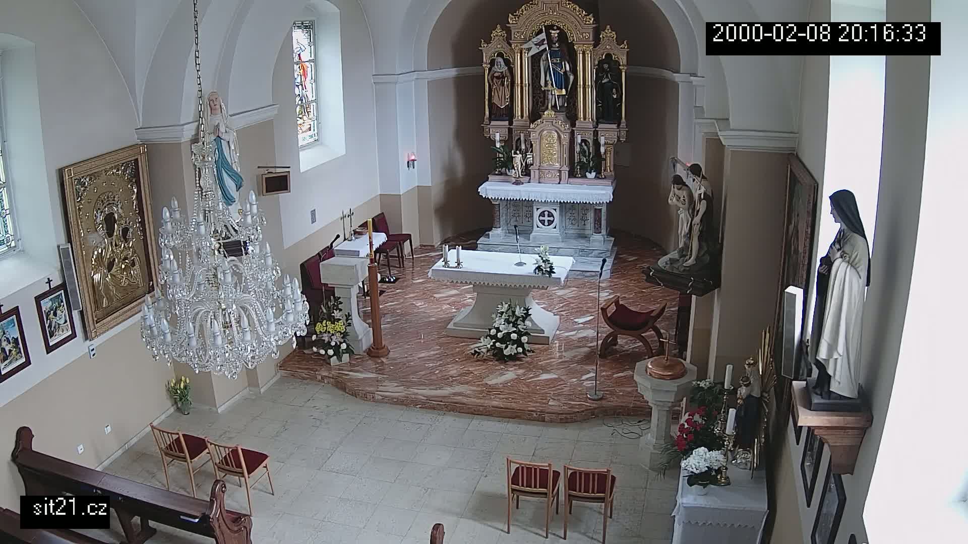 A church interior with a marble floor, an ornate chandelier, religious statues, and floral arrangements on the altar and side tables.