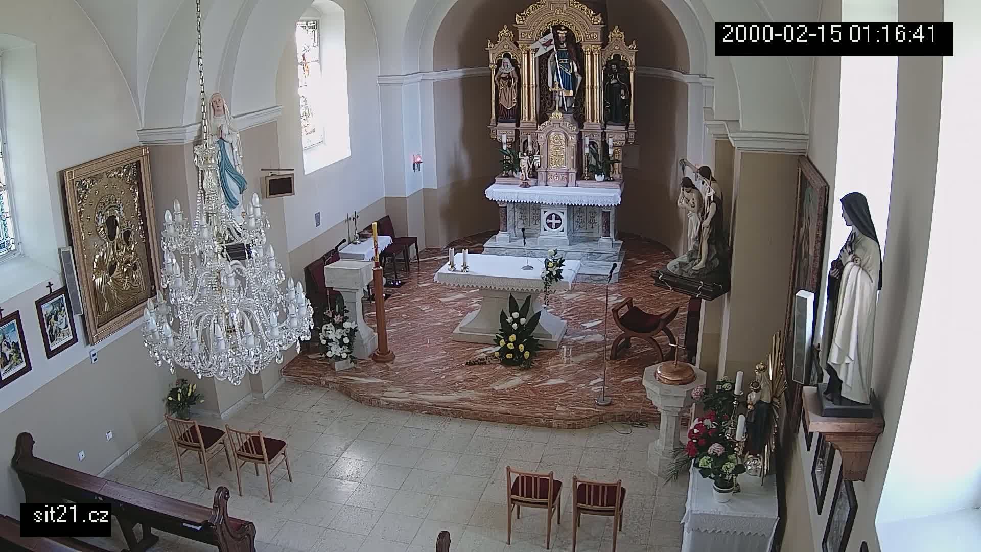 A high-angle indoor view of a church interior shows a marble floor, an altar with floral arrangements, statues, paintings, a large chandelier, and several chairs.