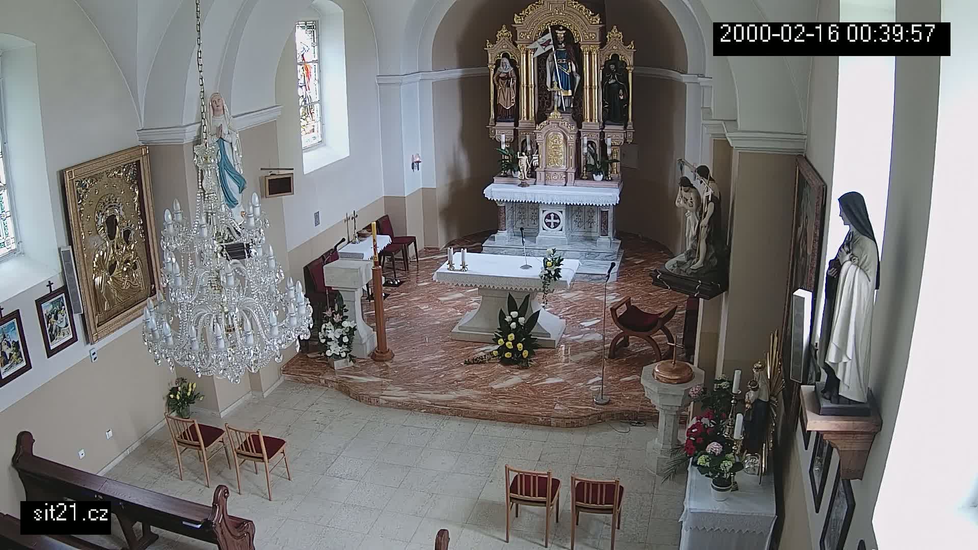 An indoor view of a church sanctuary shows an altar with floral arrangements, statues, a large chandelier, and several chairs.