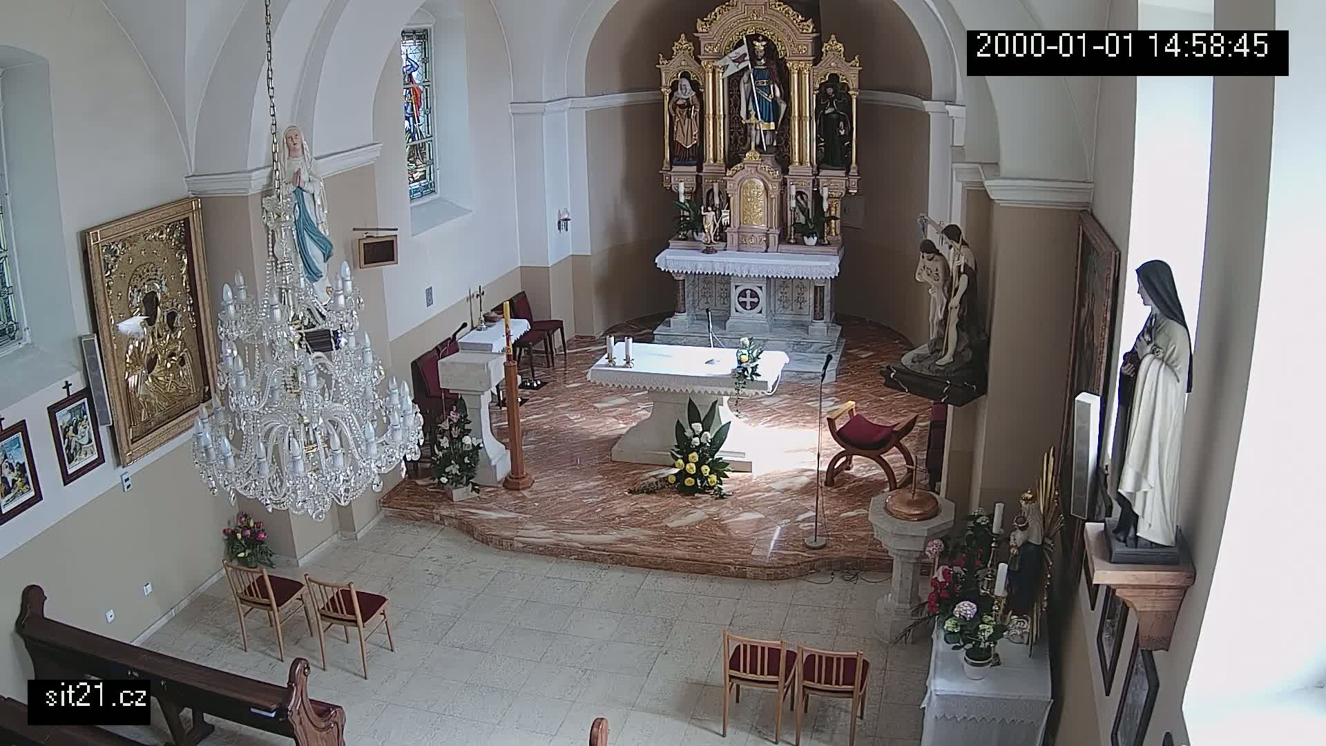A church interior, brightly lit, shows an altar with floral arrangements, statues, and a large crystal chandelier.