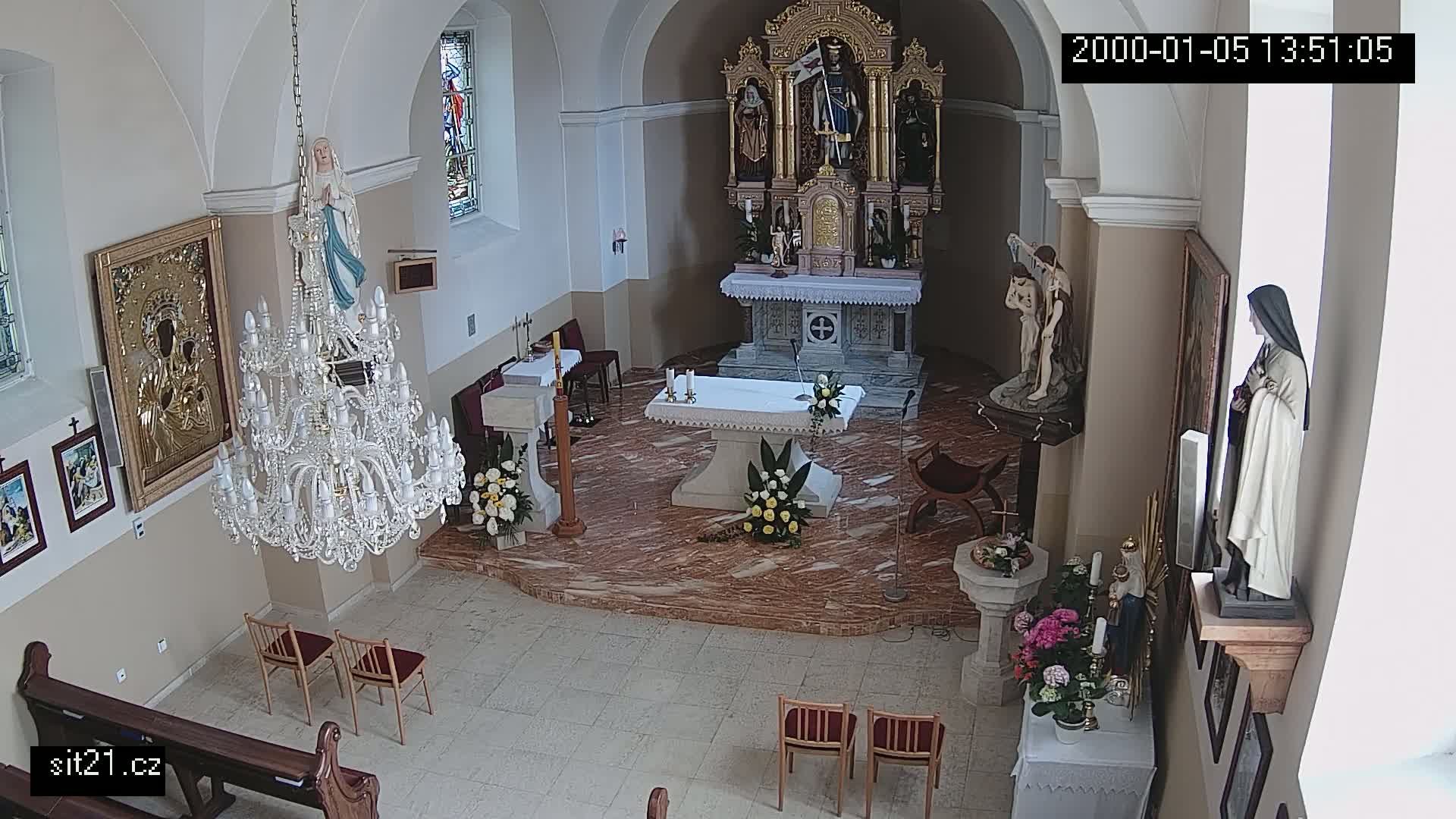 An indoor view of a church sanctuary, showing an altar with floral arrangements, religious statues, a large chandelier, and several pews.