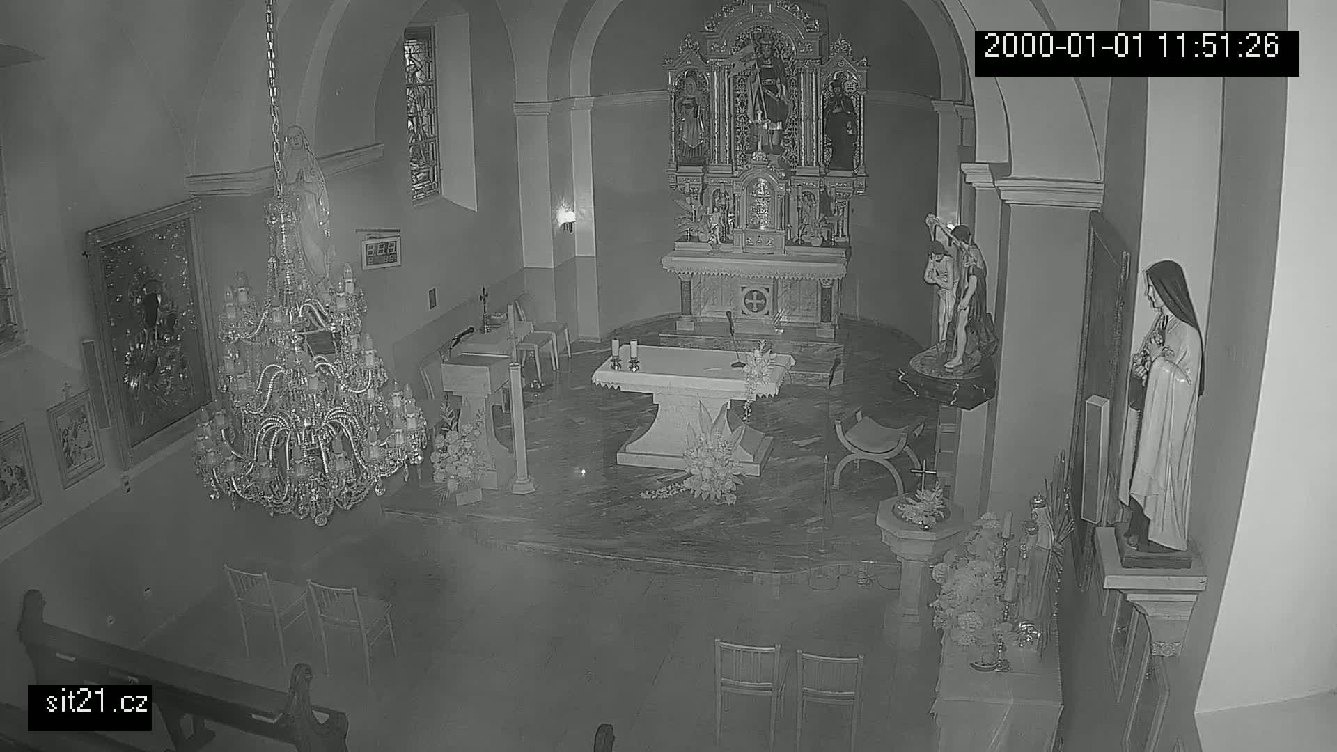 A high-angle, black and white nighttime view of the interior of a church, showing an altar, several statues, a large chandelier, and empty chairs.