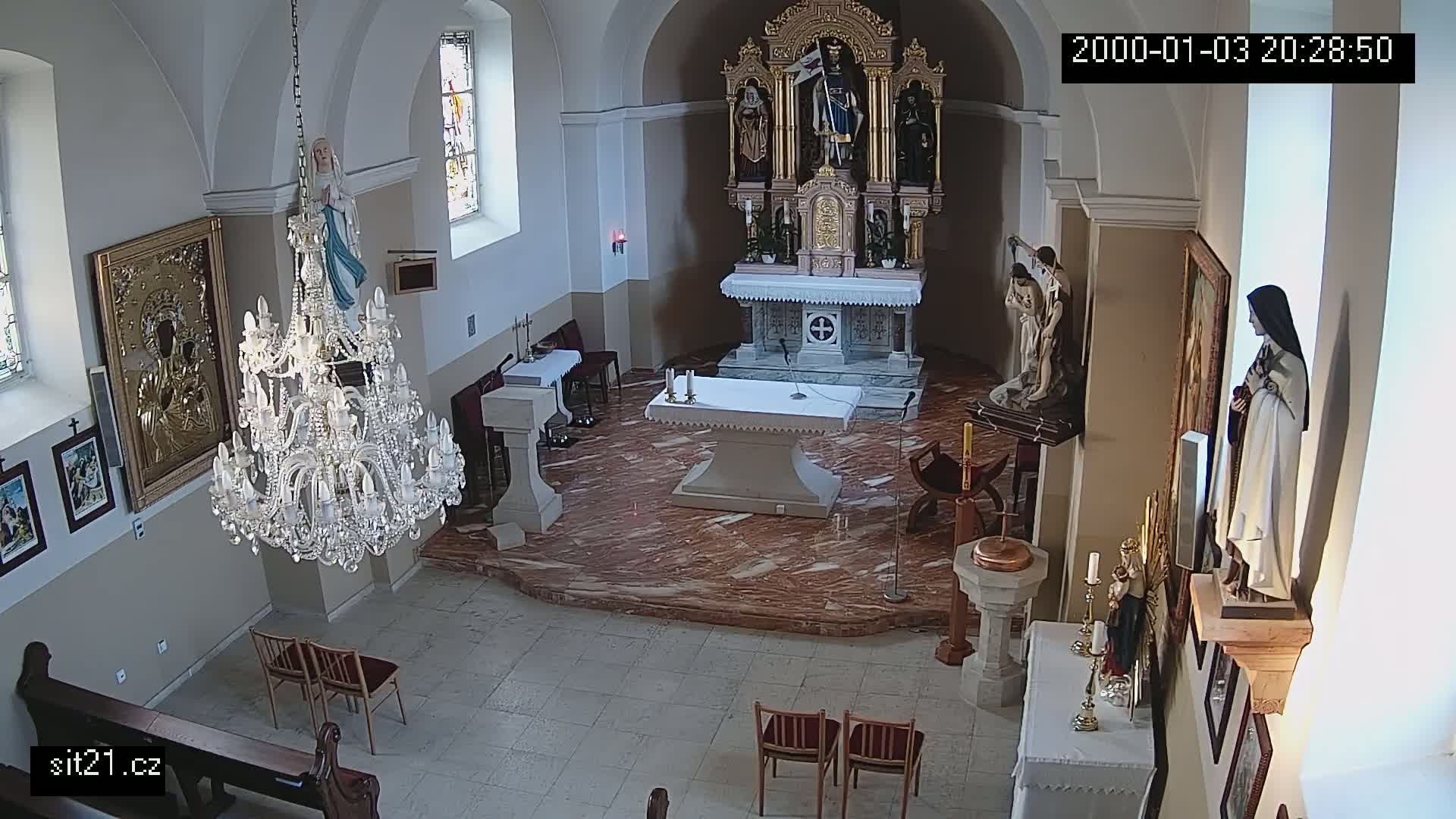 An indoor view of a church sanctuary shows an altar, several religious statues and paintings, a large crystal chandelier, and a few empty chairs.