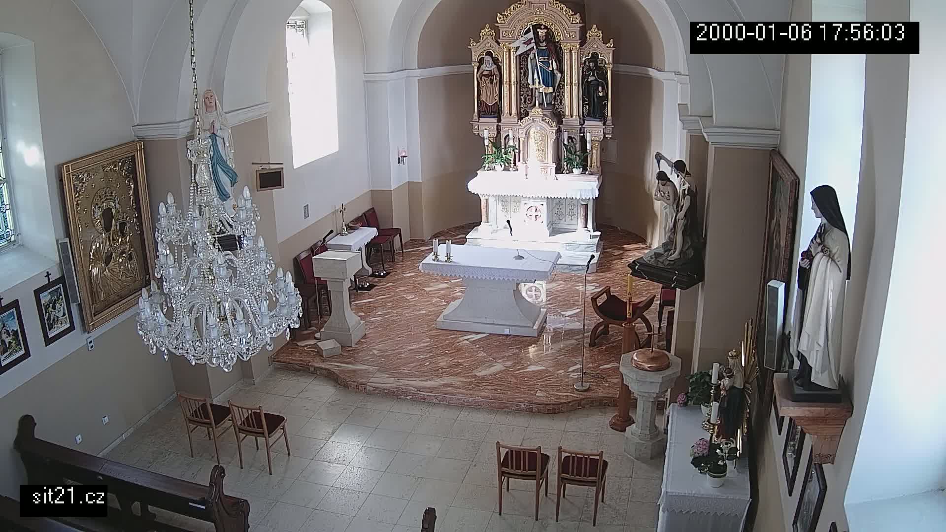 A church interior with marble floors, an altar, several statues, a large chandelier, and wooden chairs is shown in an overhead shot.