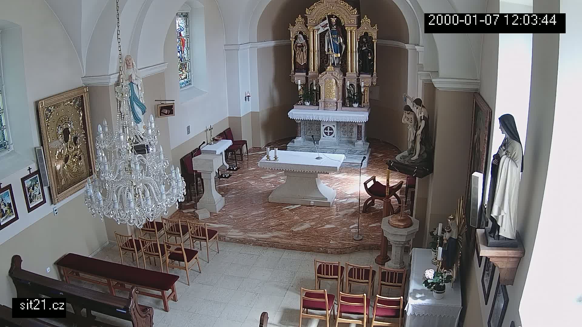 An indoor view of a church sanctuary with marble flooring, showing an altar, religious statues, a large chandelier, and rows of empty pews.