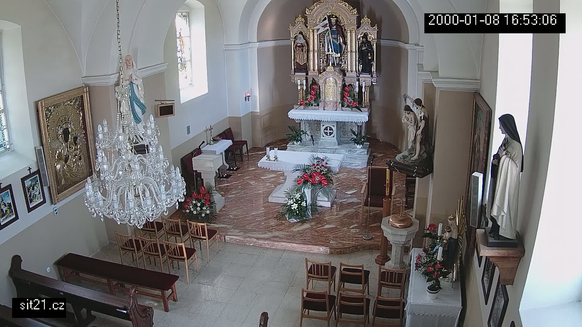 An indoor view of a church sanctuary, featuring an altar with floral arrangements, statues, a large crystal chandelier, and rows of empty chairs.