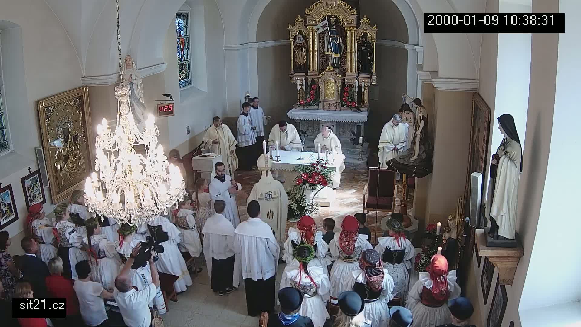 A large group of people in traditional clothing are gathered inside a church during a religious ceremony, with several priests at the altar.