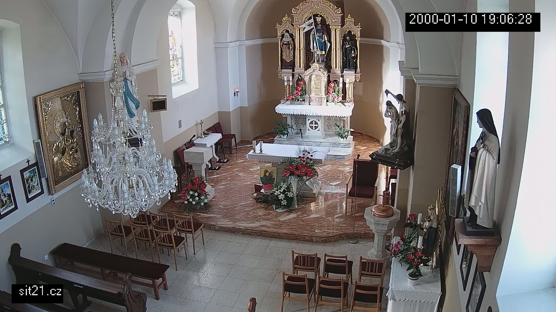 A church interior with a marble floor, an ornate altar, several statues, a large crystal chandelier, and floral arrangements.