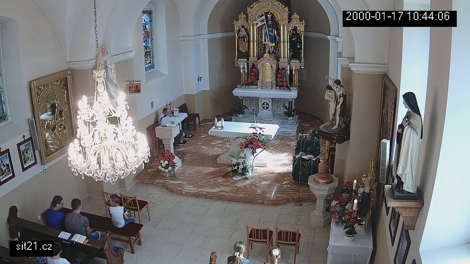 A church interior, brightly lit by sunlight streaming through windows, shows several people sitting in pews while others participate in a religious service near the altar.