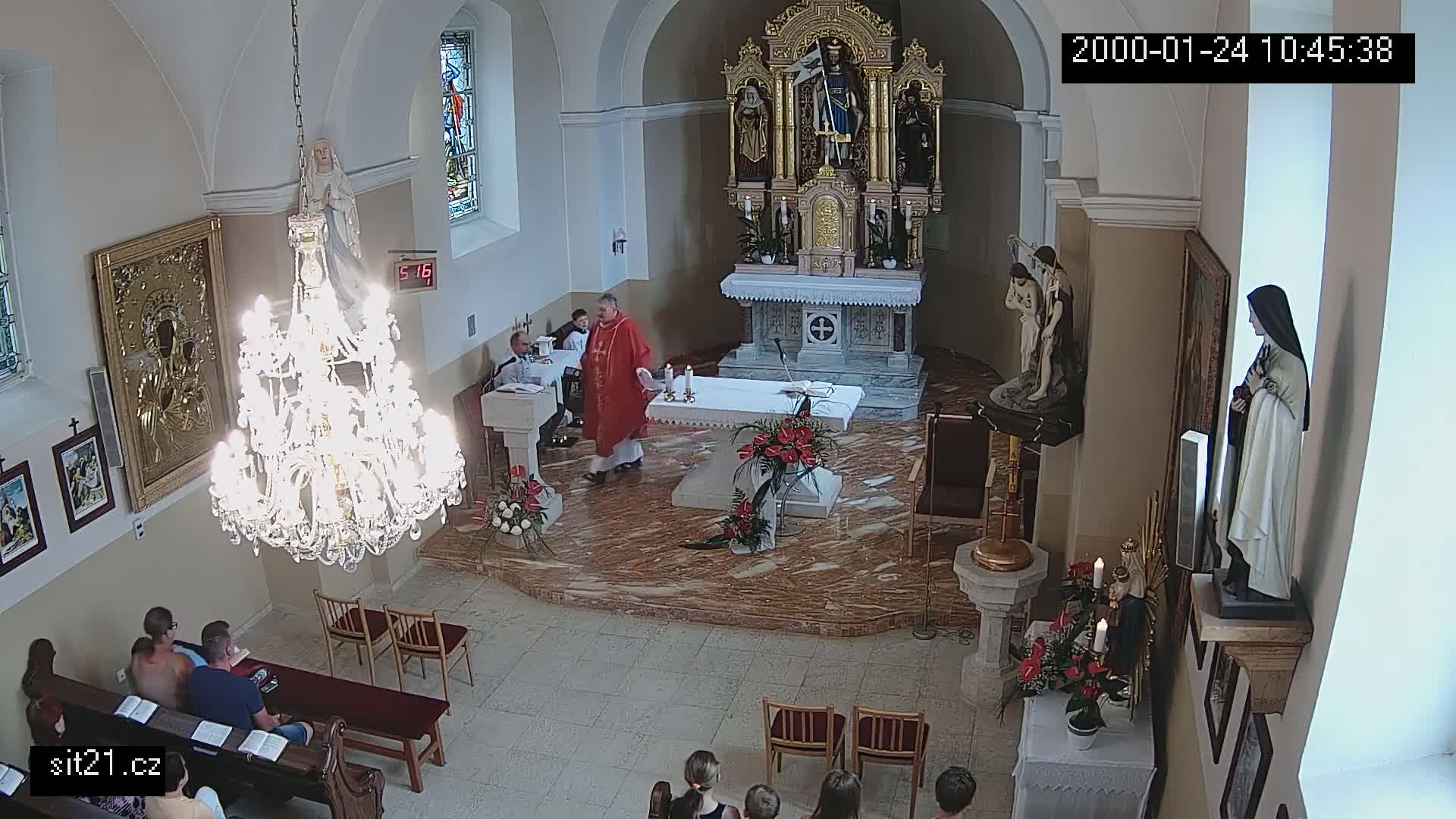 A priest in red vestments stands at an altar in a church, with several people seated in pews, while a large chandelier hangs from the ceiling.