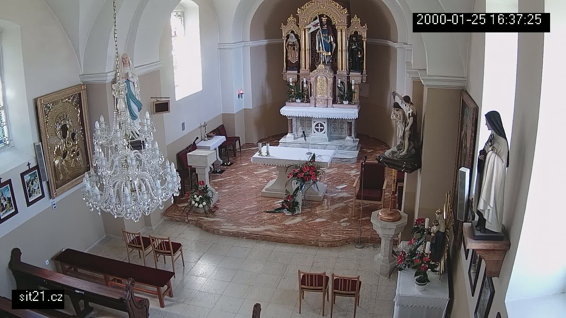 A priest in red vestments stands at an altar in a church, with several people seated in pews, while a large chandelier hangs from the ceiling.