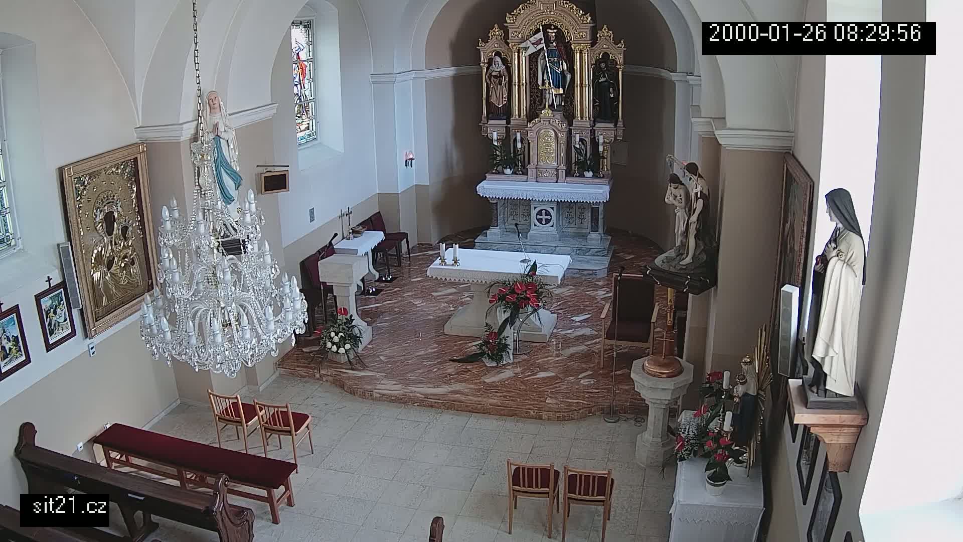 An interior view of a church sanctuary shows an altar, religious statues, a large crystal chandelier, and several rows of empty chairs.
