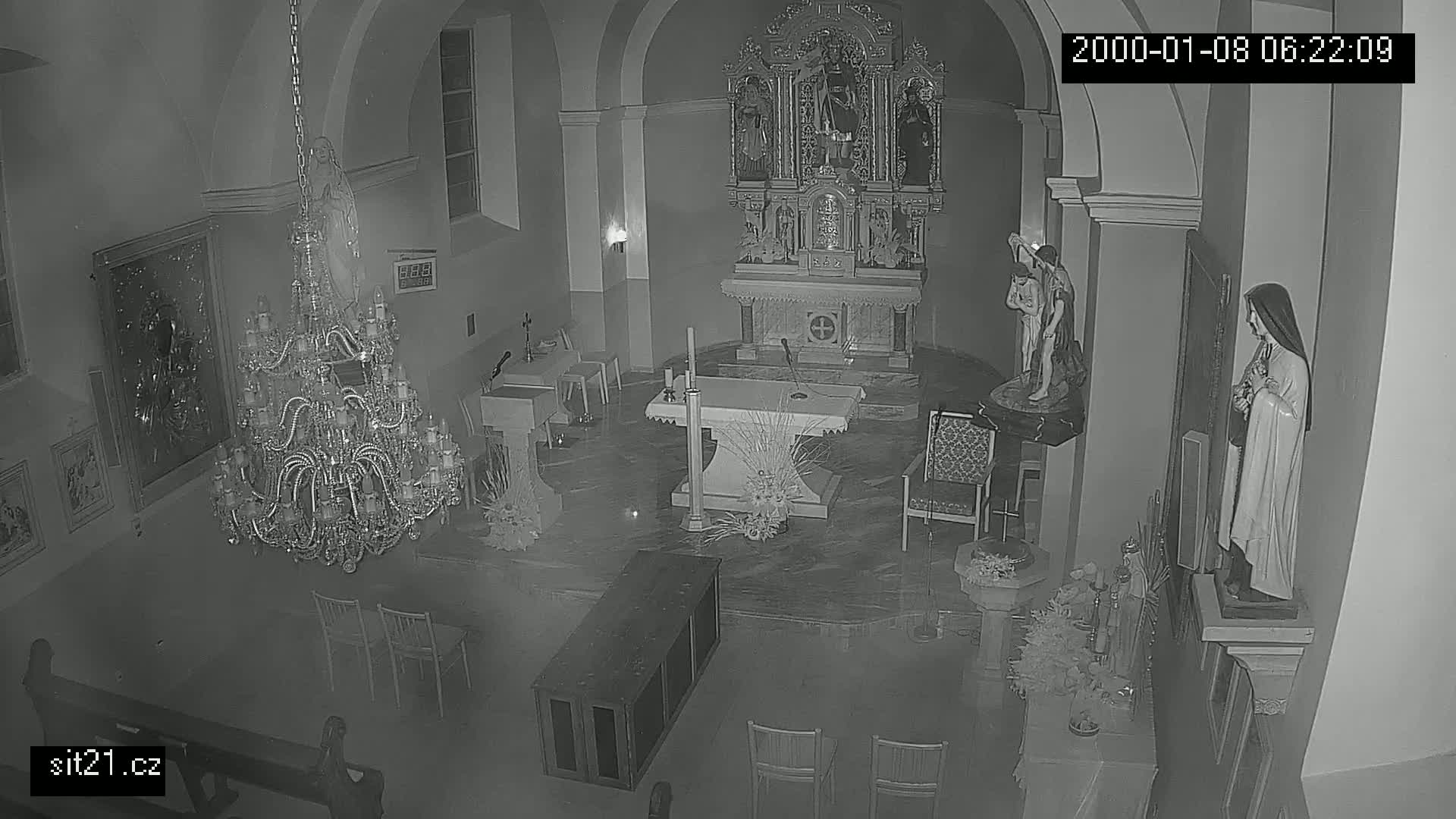 A dimly lit church interior at night shows an altar, several statues, a large chandelier, and empty pews.