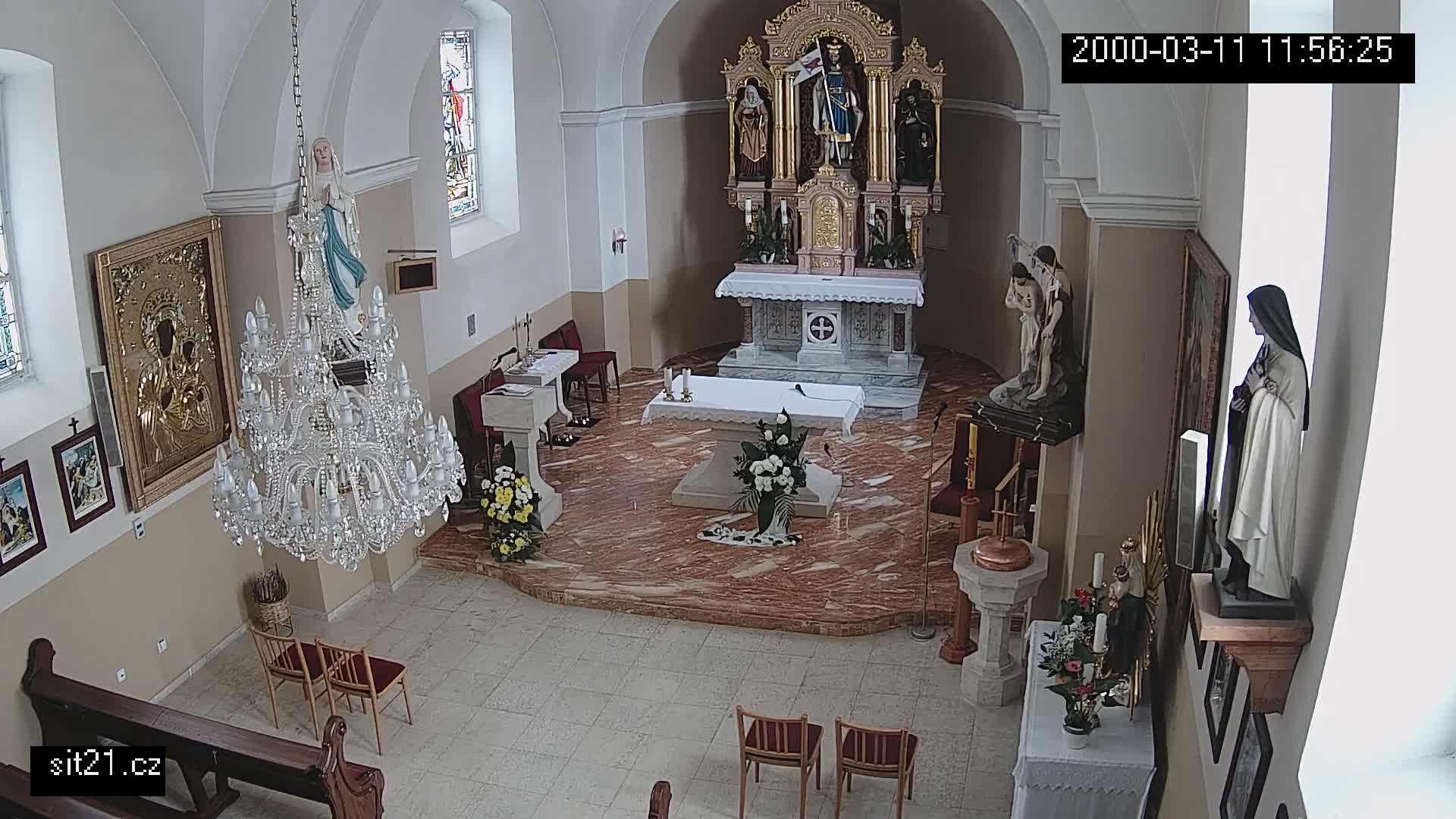 The image captures a wide interior view of a church, featuring an ornate main altar with an altarpiece, multiple religious statues, a large crystal chandelier, stained glass windows, and various religious furnishings in a spacious setting.