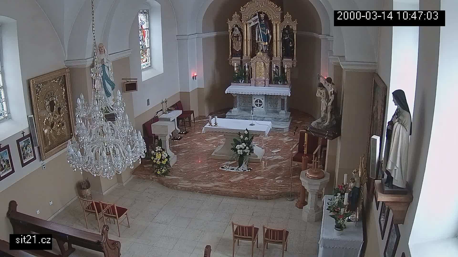 The image displays the ornate interior of a church, featuring a grand altar with a sculpted reredos, several religious statues including one of the Virgin Mary, a large crystal chandelier, and stained glass windows.