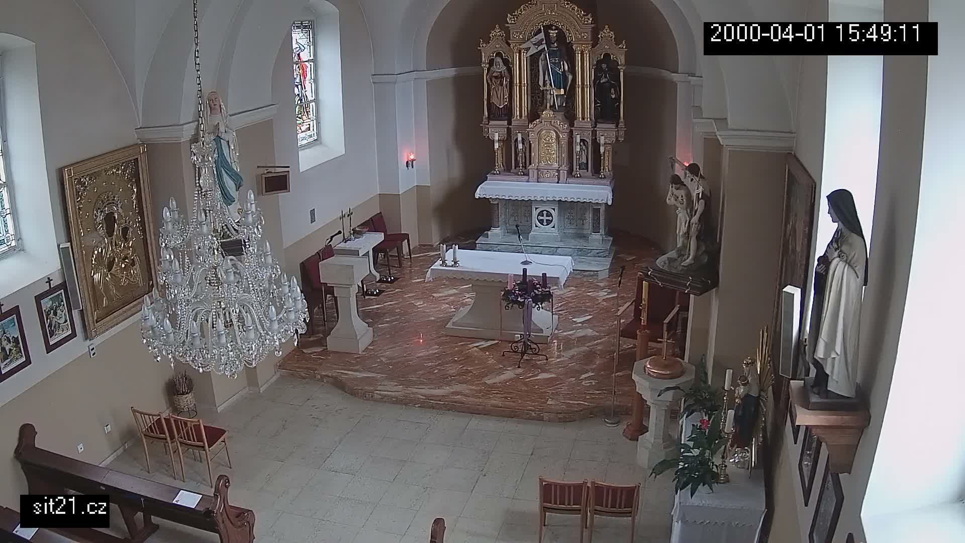 An ornate crystal chandelier hangs prominently in a church interior, which features a decorated main altar with multiple statues, a foreground altar with candles and flowers, and stained glass windows.