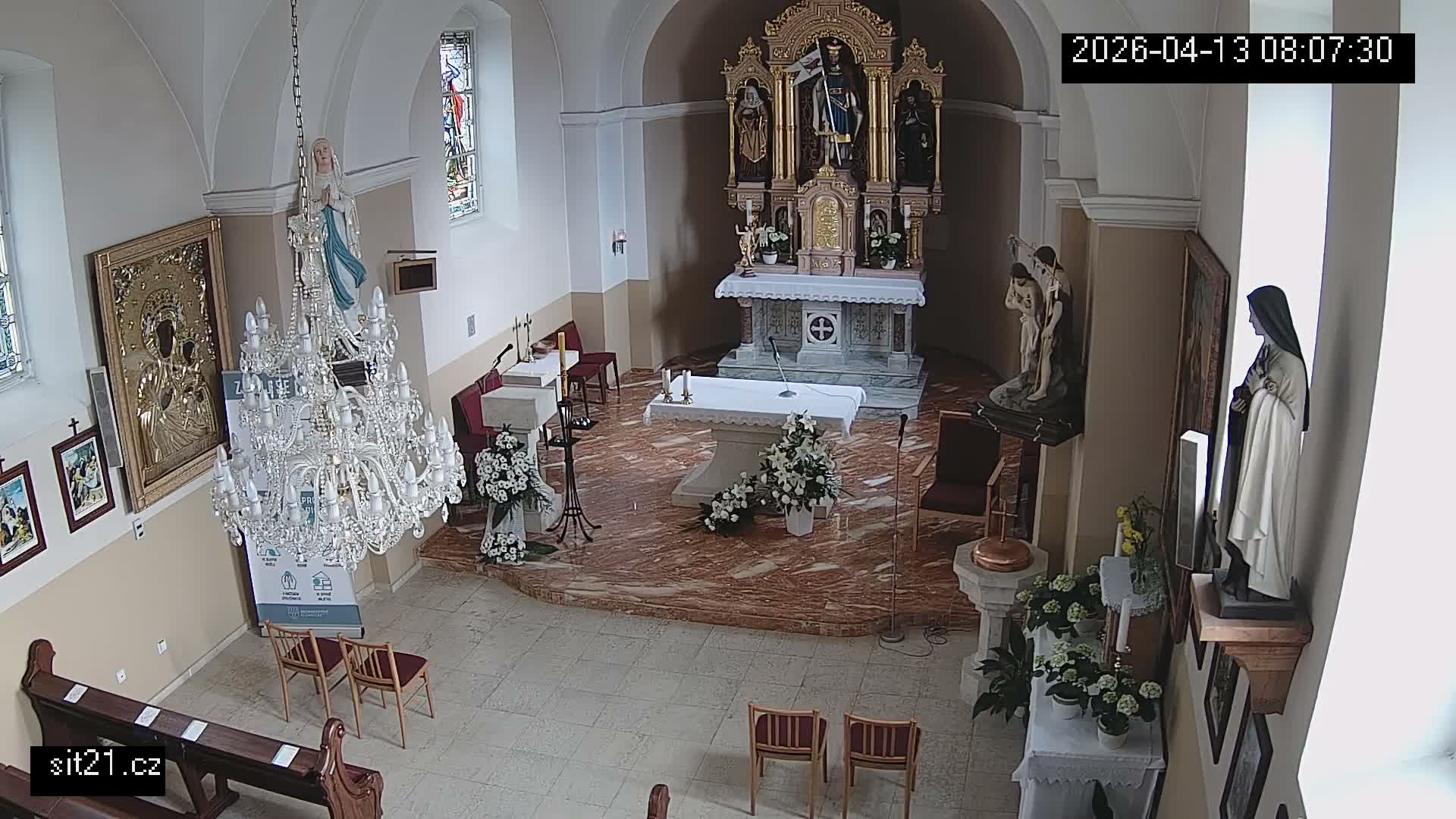 An ornate crystal chandelier hangs prominently in a church interior, which features a decorated main altar with multiple statues, a foreground altar with candles and flowers, and stained glass windows.