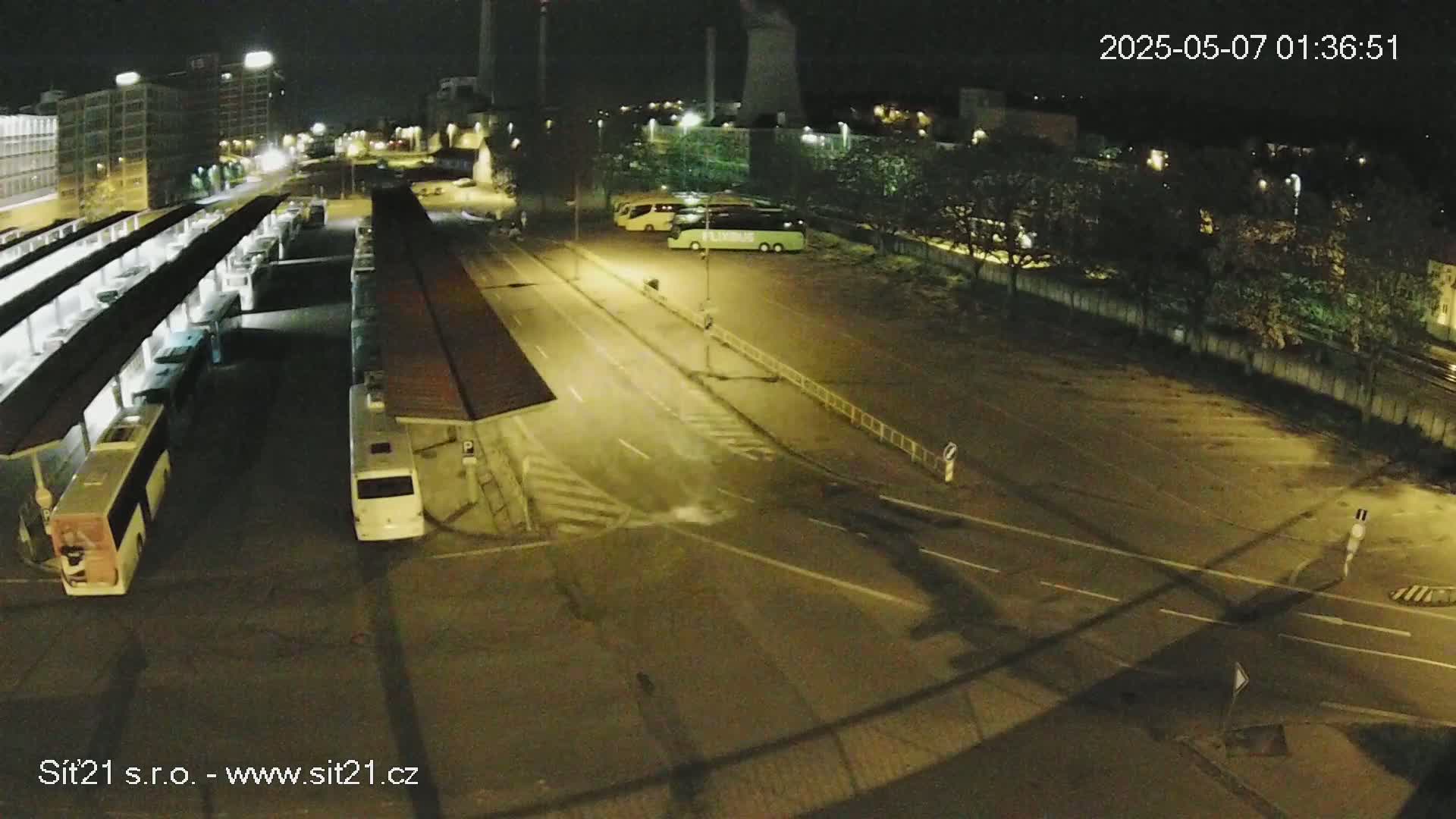 A nighttime aerial view shows a bus depot with several buses parked under shelters, alongside a roadway intersecting with another road where there are additional parked buses.
