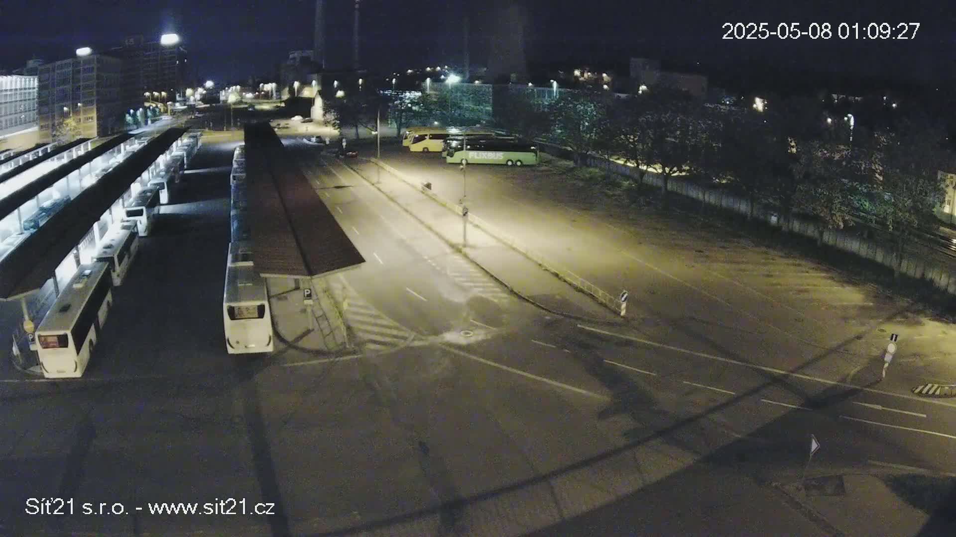 A nighttime, high-angle view shows a bus station with several buses parked under shelters, adjacent to a roadway and a larger parking lot containing additional buses.