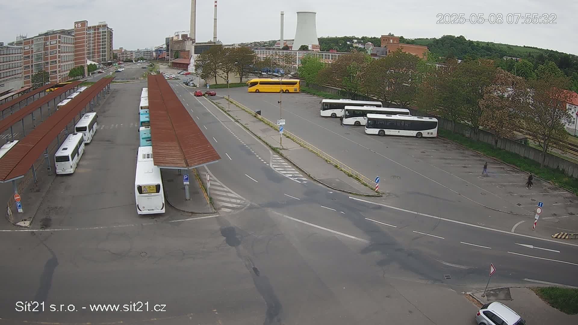 An overcast day at a bus station shows several buses parked under shelters and in a lot, with a few cars visible on surrounding roads.