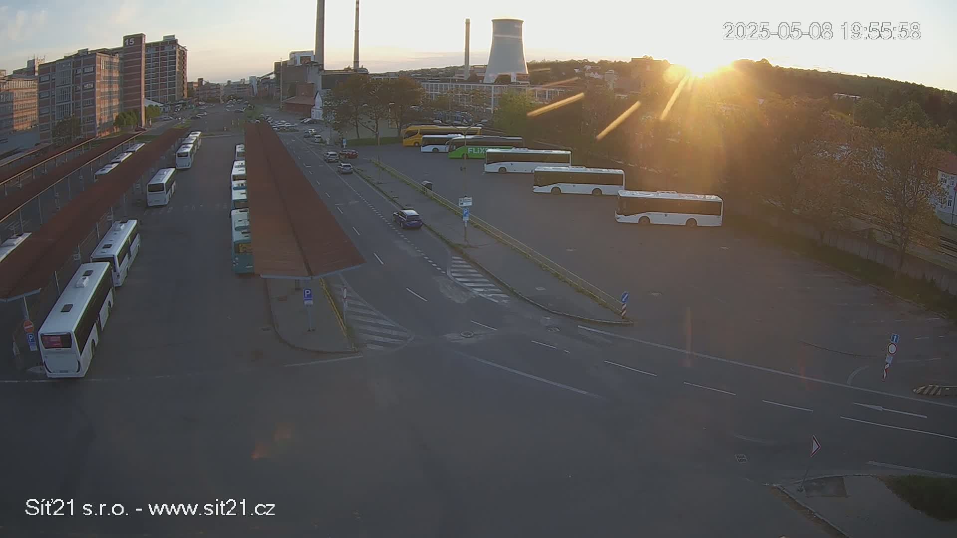 A high-angle, long shot shows a bus station with numerous buses parked at sunset, with a factory and cooling tower in the background.