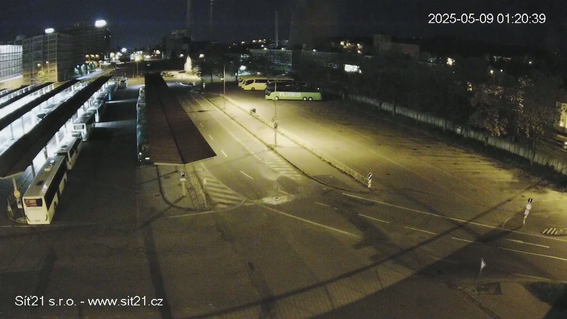At night, a mostly empty bus station with several buses parked under covered areas and a few more parked in an adjacent lot is illuminated by artificial lights.