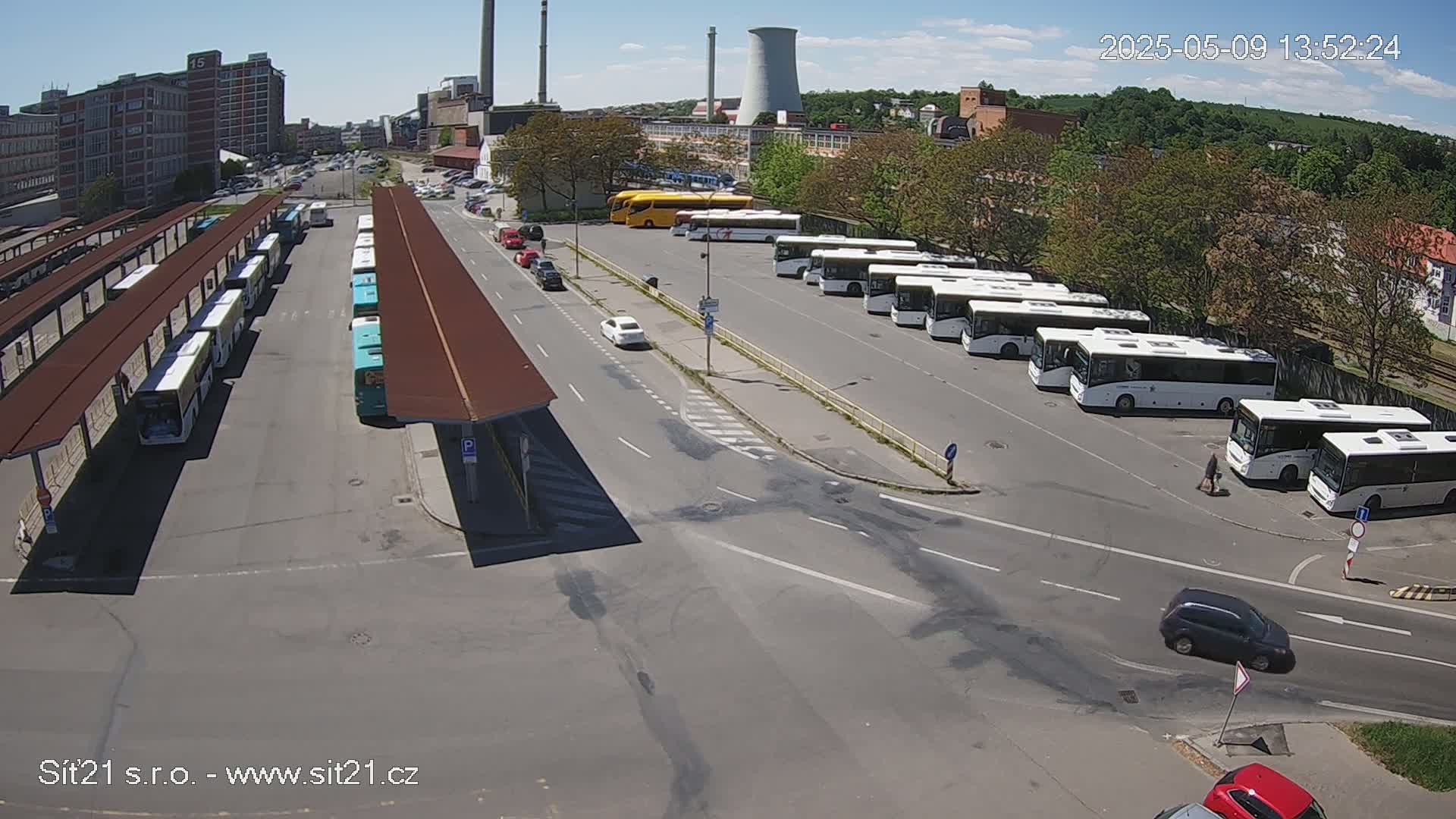 Under a sunny sky, a bus depot shows numerous buses parked at covered and uncovered areas, with several cars visible on nearby roads.