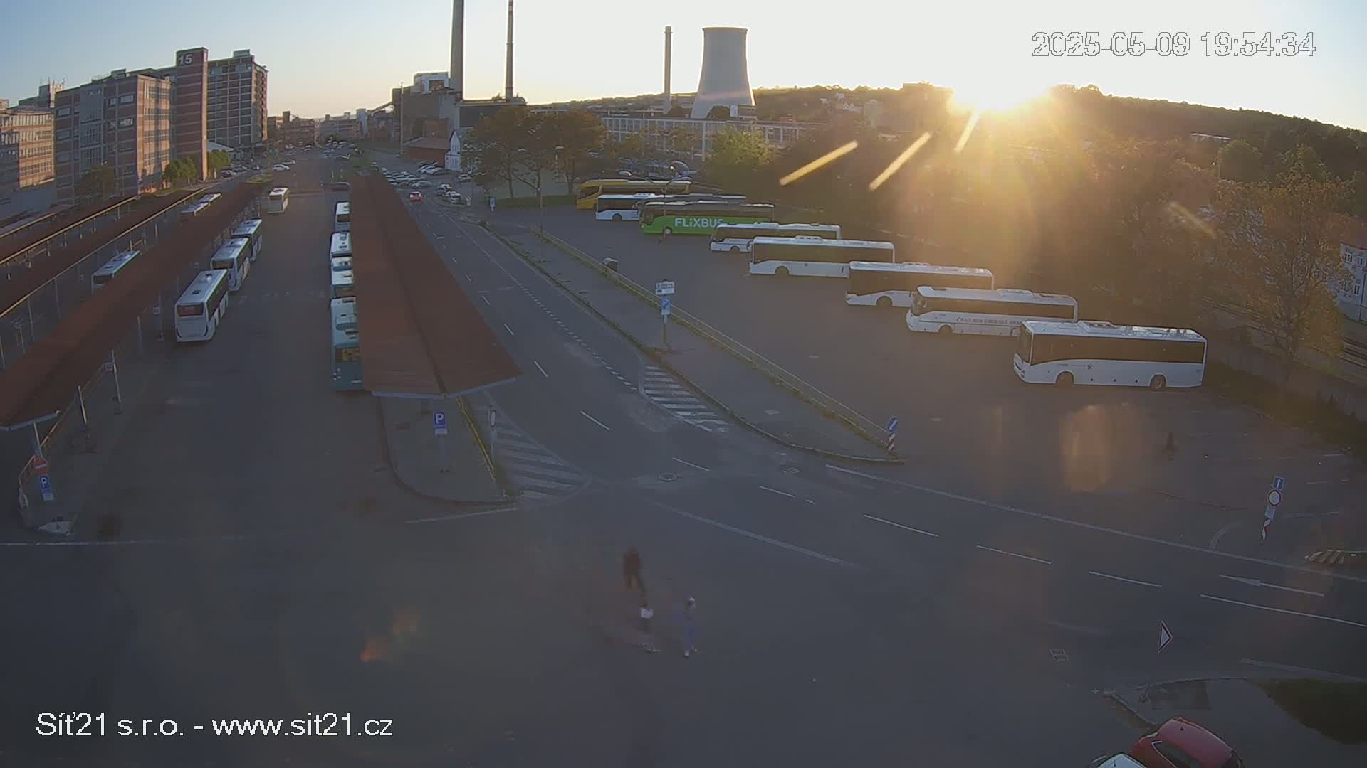 A high-angle, long shot shows a bus station with numerous buses parked at the platforms under the evening sun, with industrial buildings and trees visible in the background.