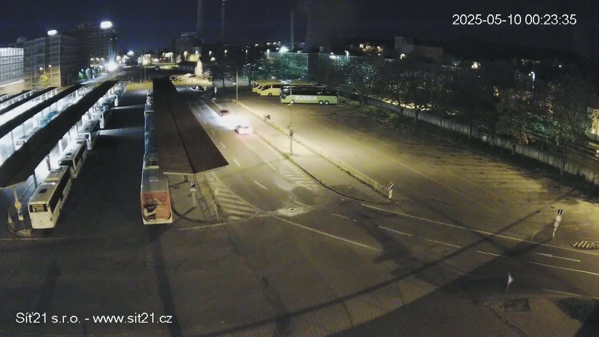 A nighttime, overhead view of a bus station with several buses parked under shelters, a car driving on the road, and a parking lot with additional buses.