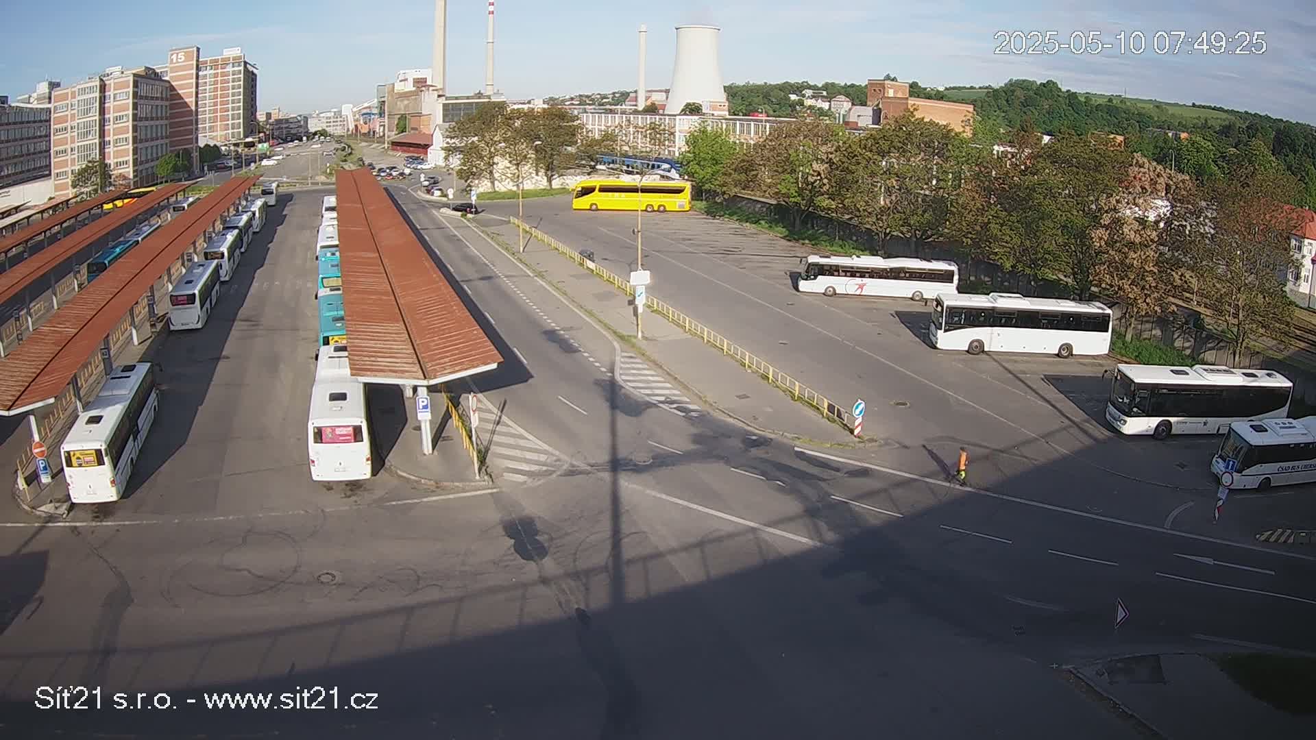 A bus station with numerous buses parked under covered shelters, along with several more buses parked in a nearby lot on a sunny day.