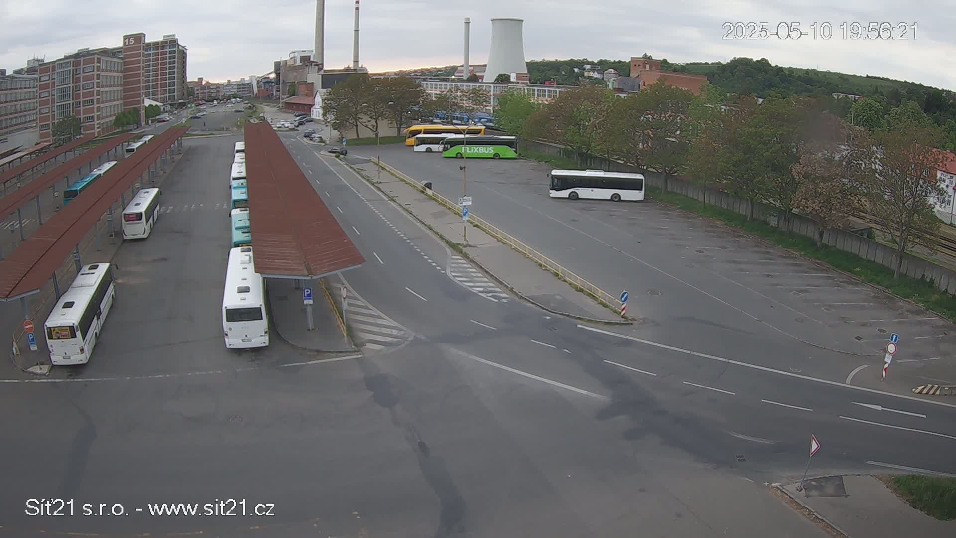 An overcast day shows a bus station with several buses parked at covered stops and in a nearby lot, near industrial buildings and trees.