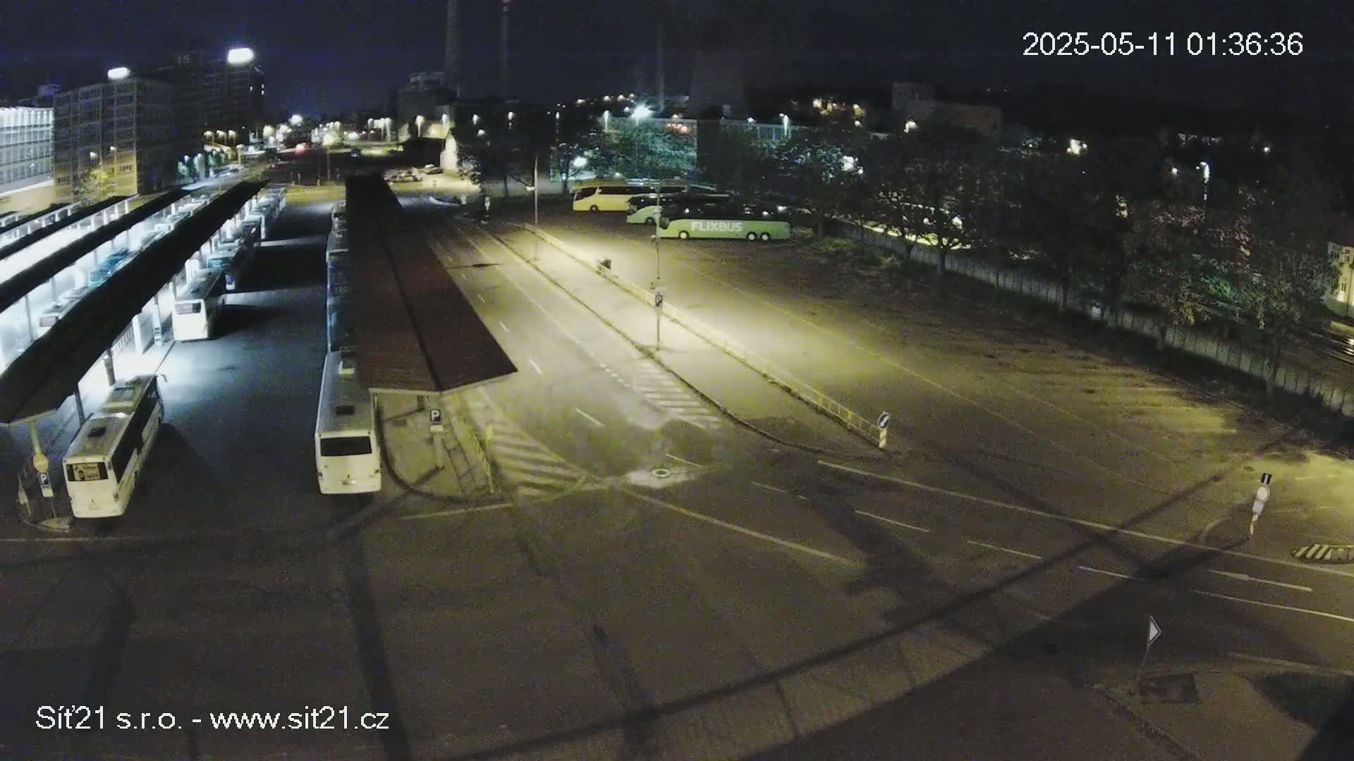 At night, a mostly empty bus station with several buses parked under shelters and a few more in a nearby lot is illuminated by artificial lights.