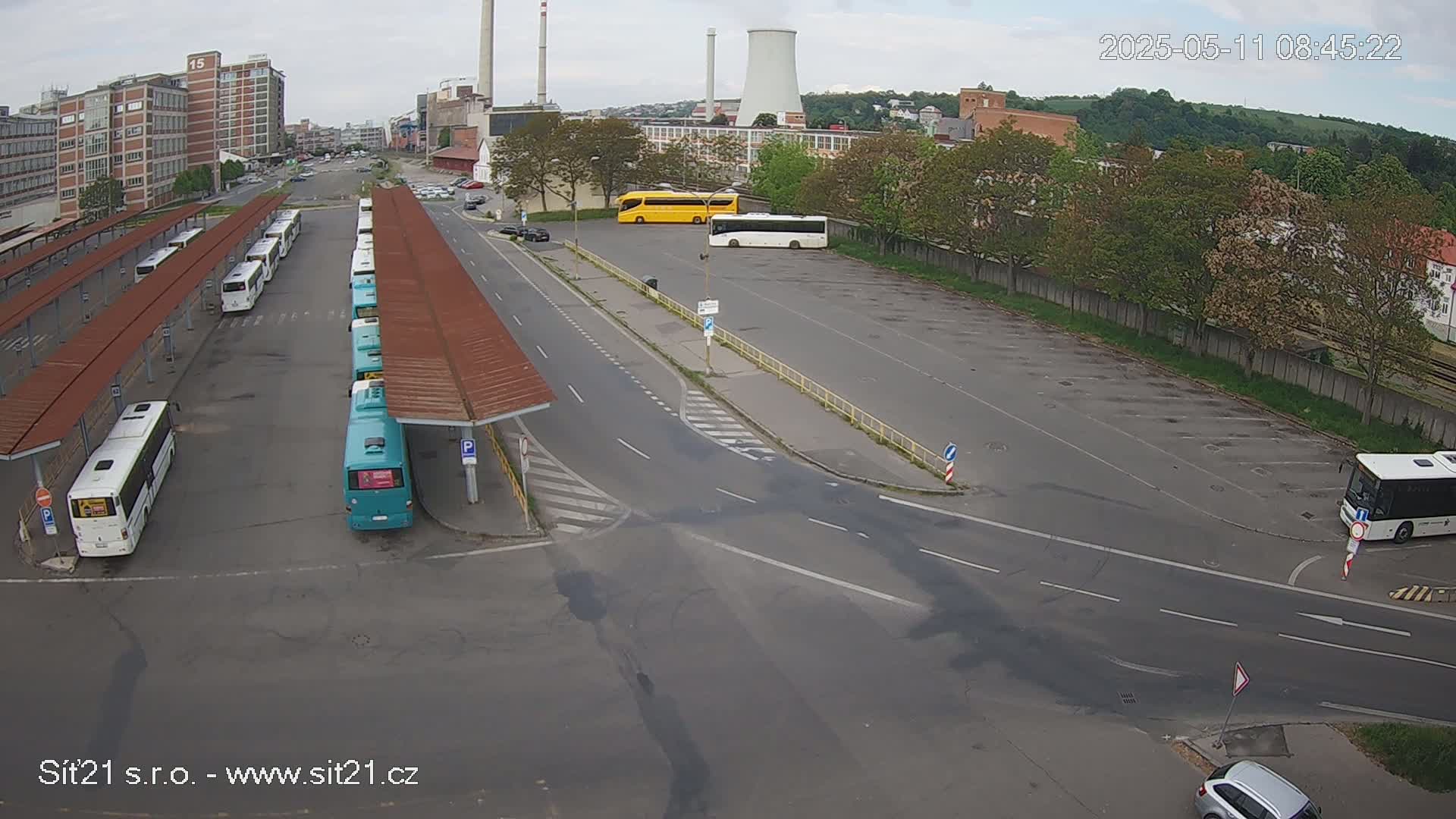 An overcast day shows a bus station with many buses parked under shelters, along with several other buses parked in an adjacent lot, and a few cars nearby.