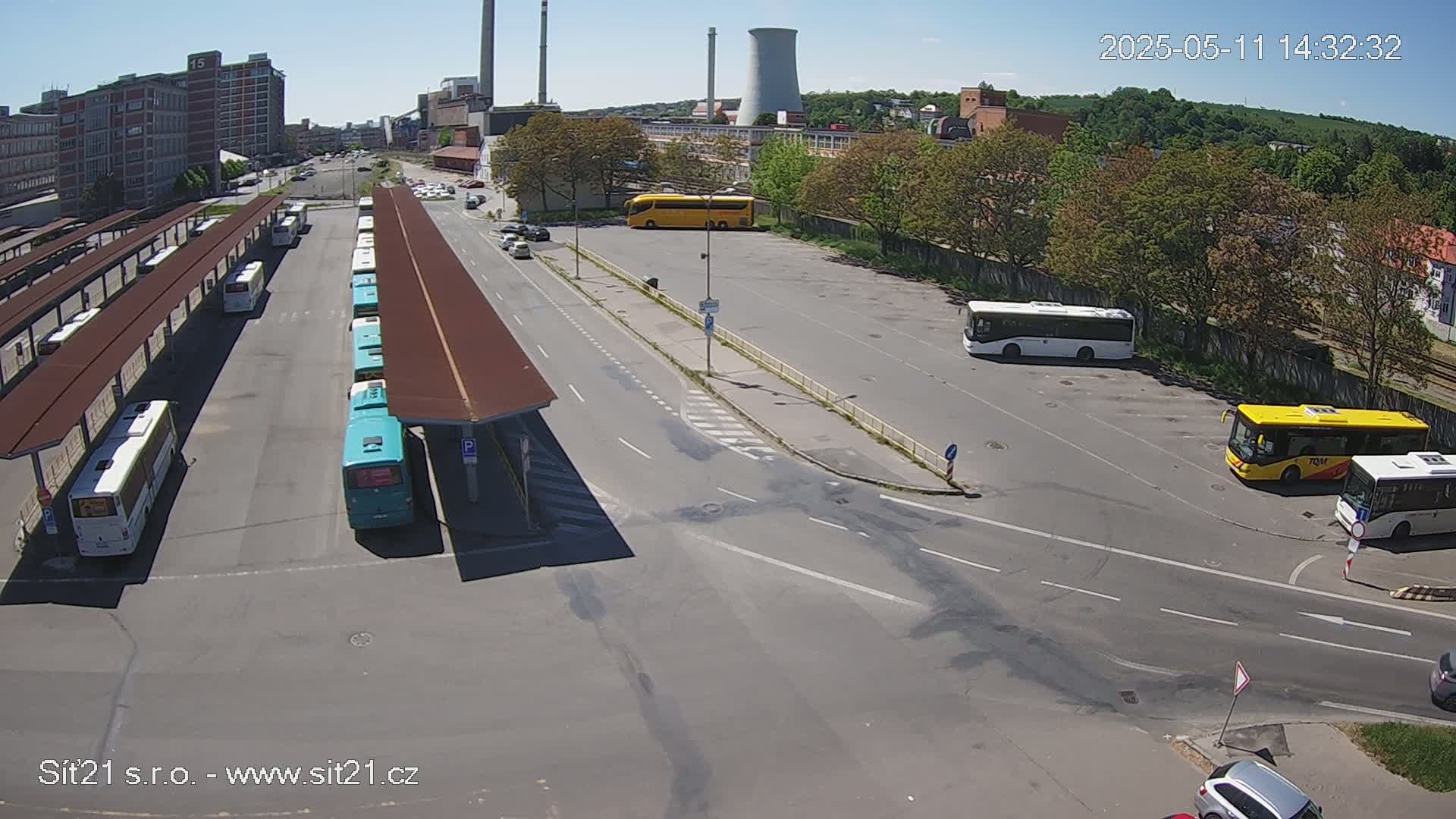 A bus station with numerous buses parked under shelters on a sunny day, with additional buses and cars in a nearby lot and on roads adjacent to industrial buildings in the background.