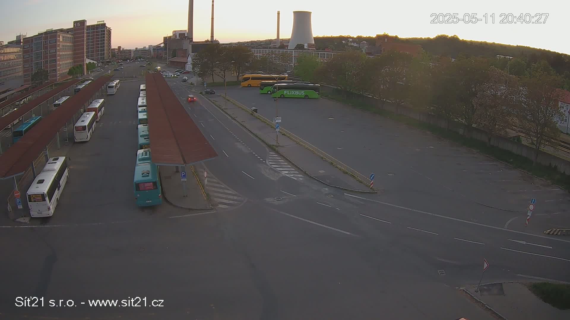 A bus station with many buses parked under covered shelters, a few more buses parked in an adjacent lot, and a road intersection, all under a dusky sky.