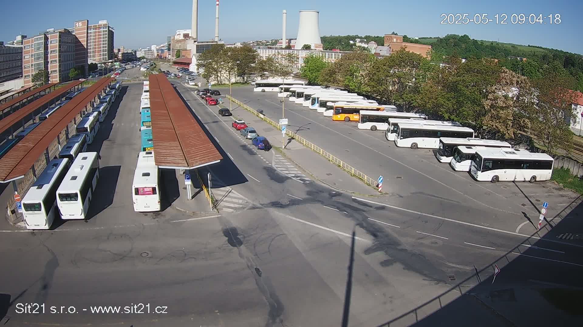 Numerous buses are parked at a bus station on a sunny day, with several cars visible on the roads nearby.