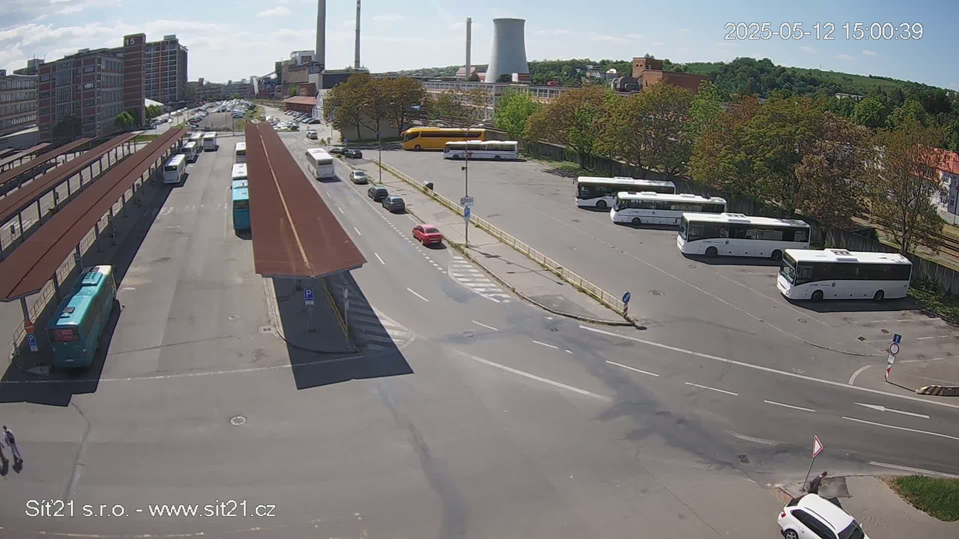 A bus station with numerous buses parked under sunny skies, adjacent to industrial buildings and trees.