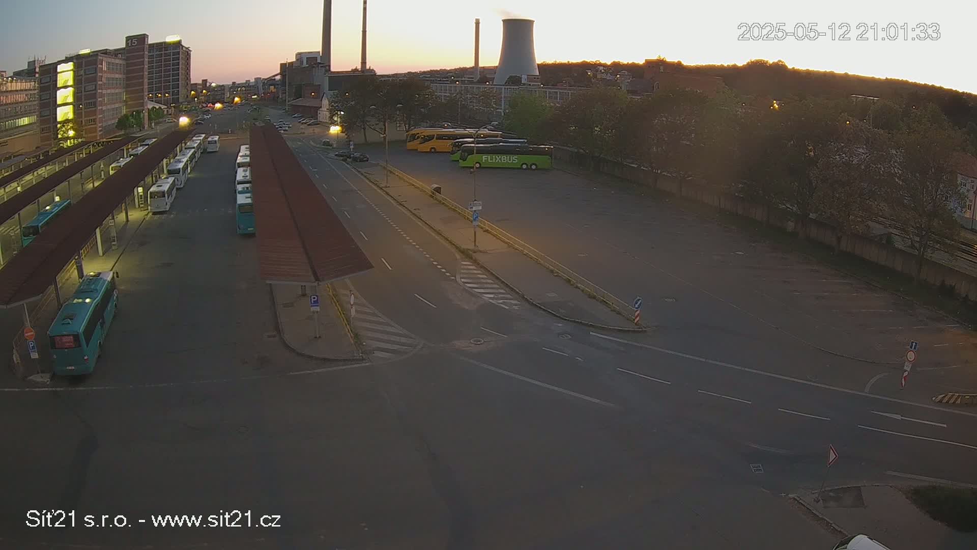A bus station with several buses parked under covered shelters, a few more buses parked in a lot across the street, and industrial buildings in the background under a twilight sky.