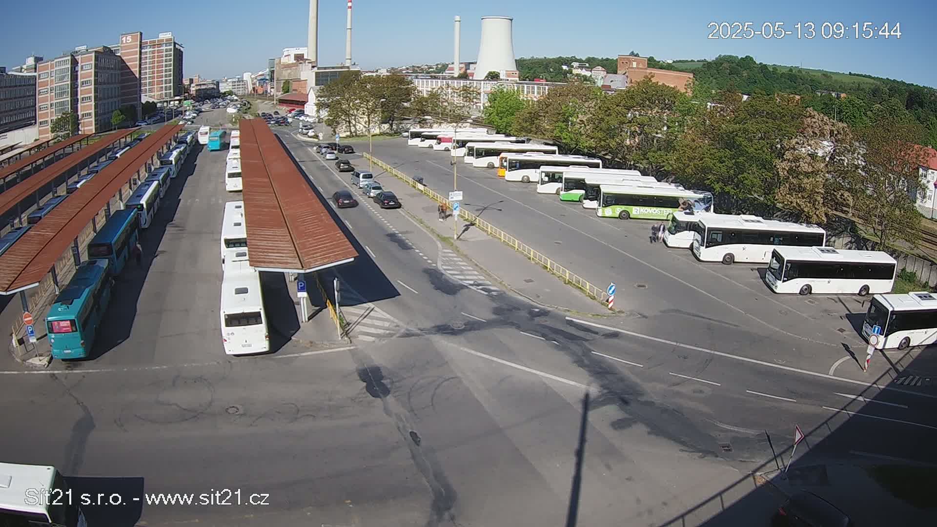 A bus station with many buses parked under covered shelters on a sunny day, near industrial buildings and a cooling tower.