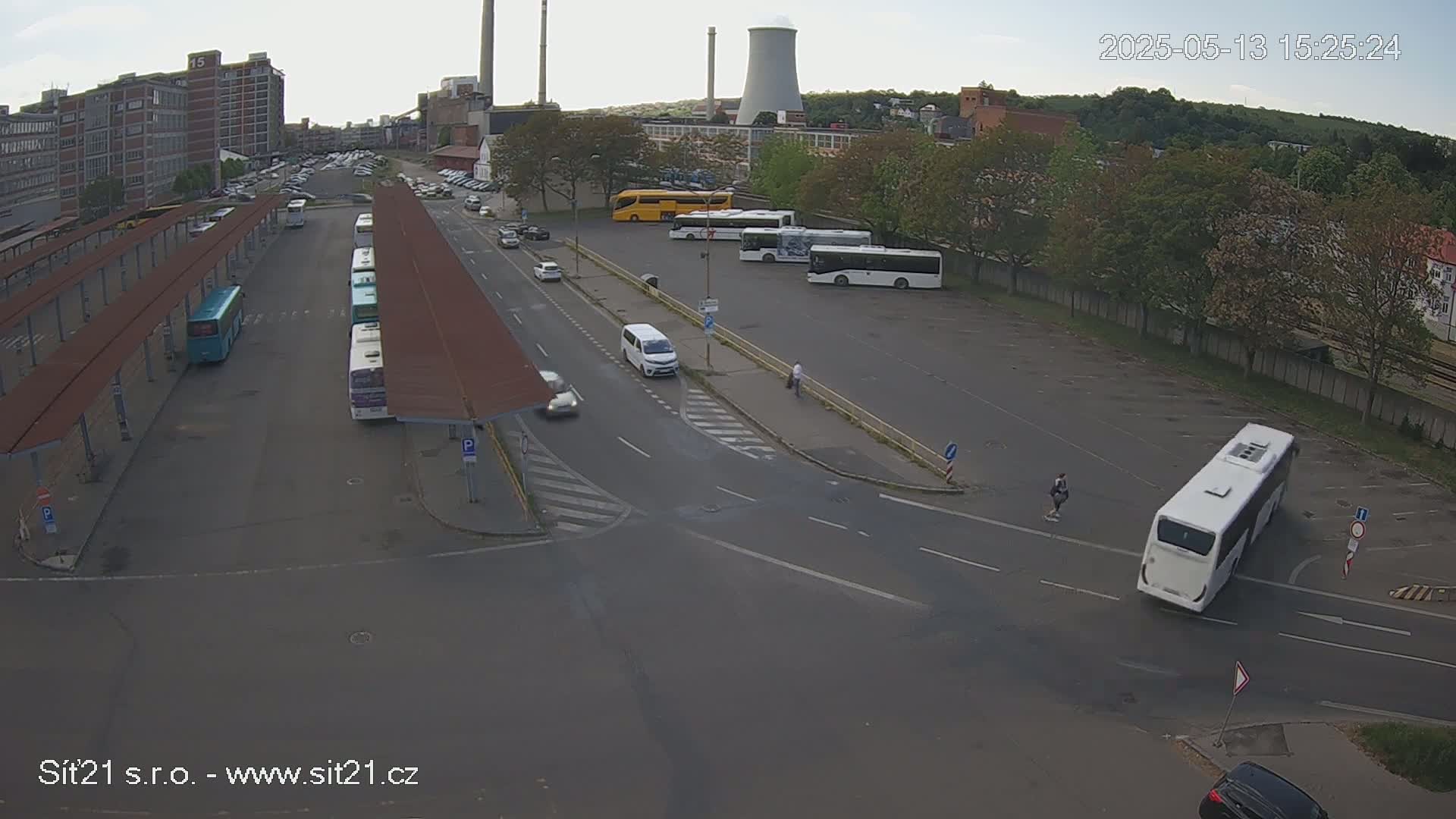 An overhead view of a bus station with several buses parked under covered areas and in a nearby lot, on a partly cloudy day.