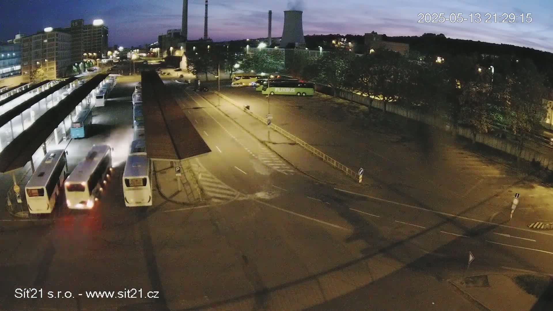 At night, under a twilight sky, several buses are parked at a bus station near a roadway and an industrial area.