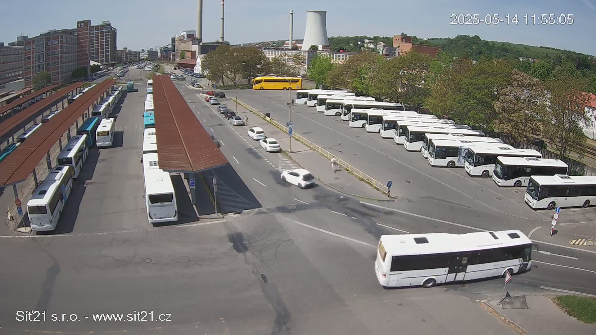A bus station with many white buses parked under covered areas, a few cars on the road, and a yellow bus parked further away on a sunny day.