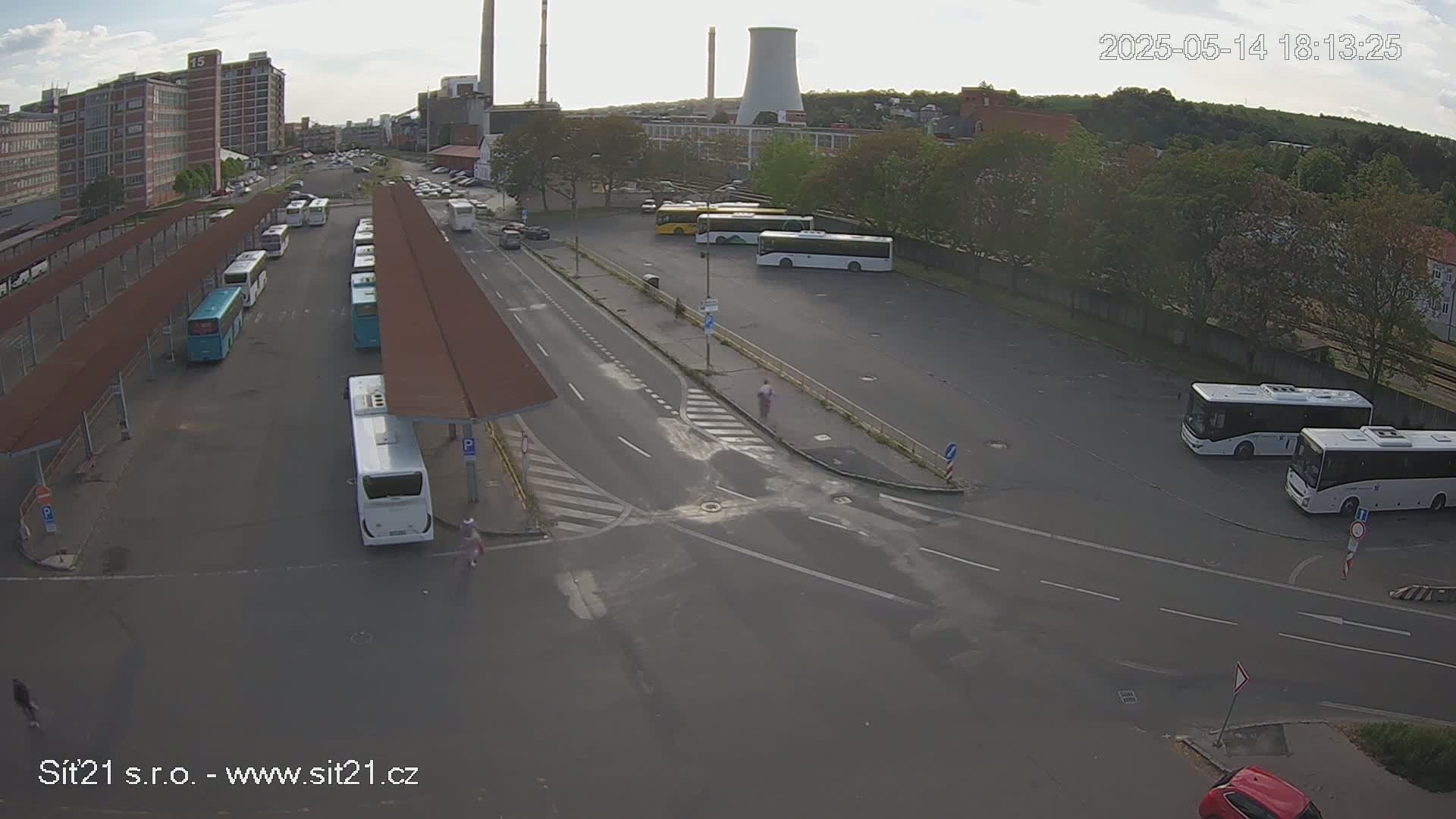 An aerial view of a bus station with numerous buses parked under shelters and in a lot, situated near industrial buildings and under a partly cloudy sky.