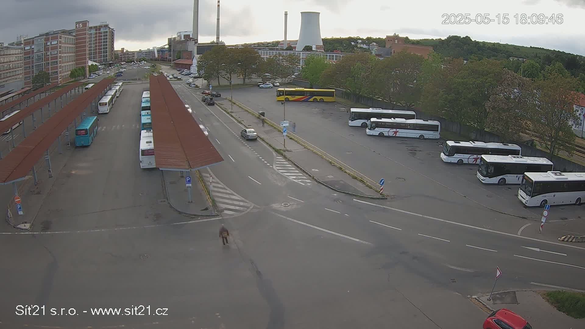 An overcast day shows a bus station with numerous buses parked under shelters and in a lot, with cars on roads surrounding the station and a cooling tower in the distance.