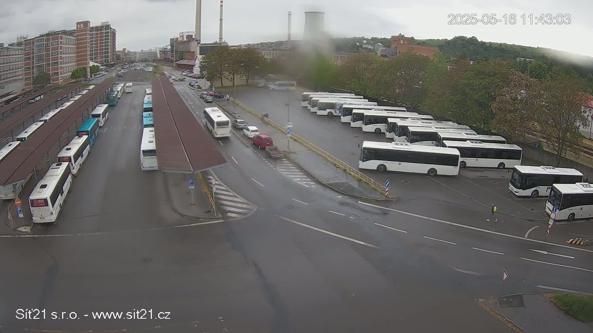 A wet bus depot with numerous buses parked under covered areas and in an open lot, surrounded by industrial buildings under an overcast sky.