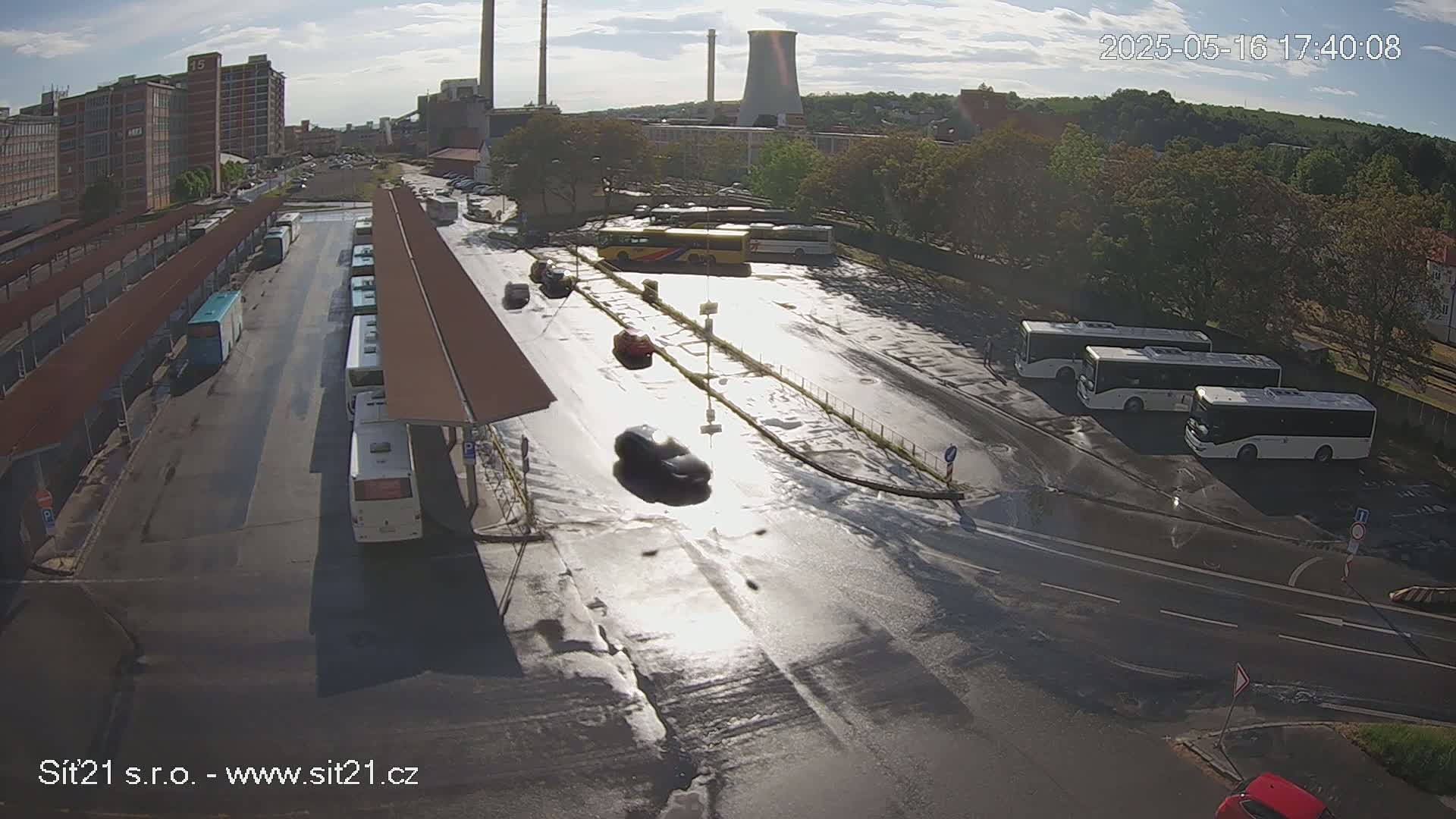 A wet bus station with several buses parked under covered shelters, cars driving on the road, and industrial buildings visible in the background under a partly sunny sky.
