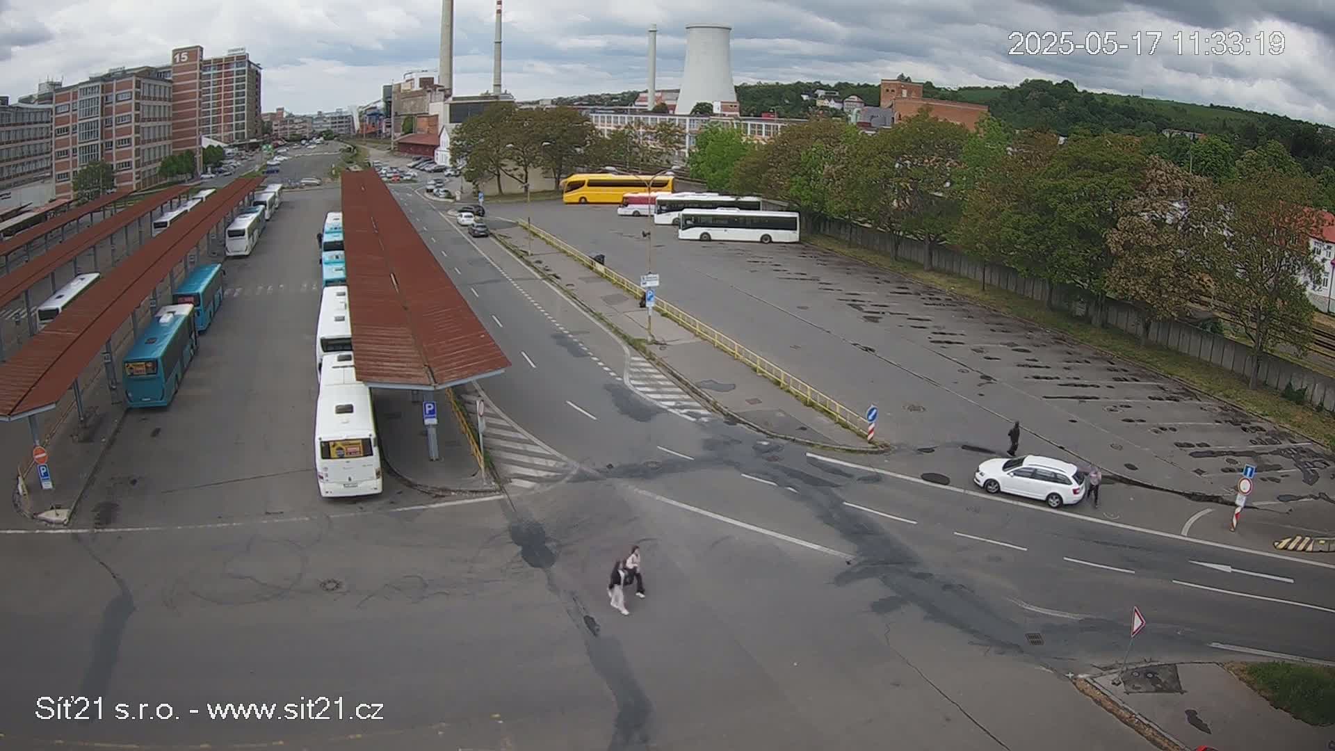 An overcast day reveals a bus station with numerous buses under shelters, a nearby empty parking lot with a white car, and industrial buildings in the background.
